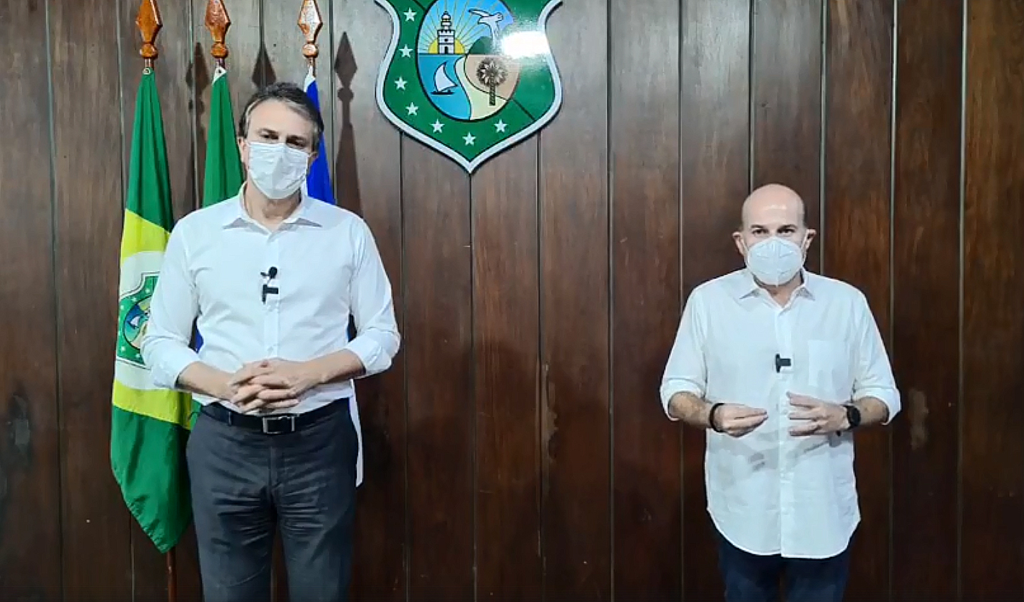 governador e prefeito lado a lado de máscara, posando para a câmera e com bandeiras ao fundo